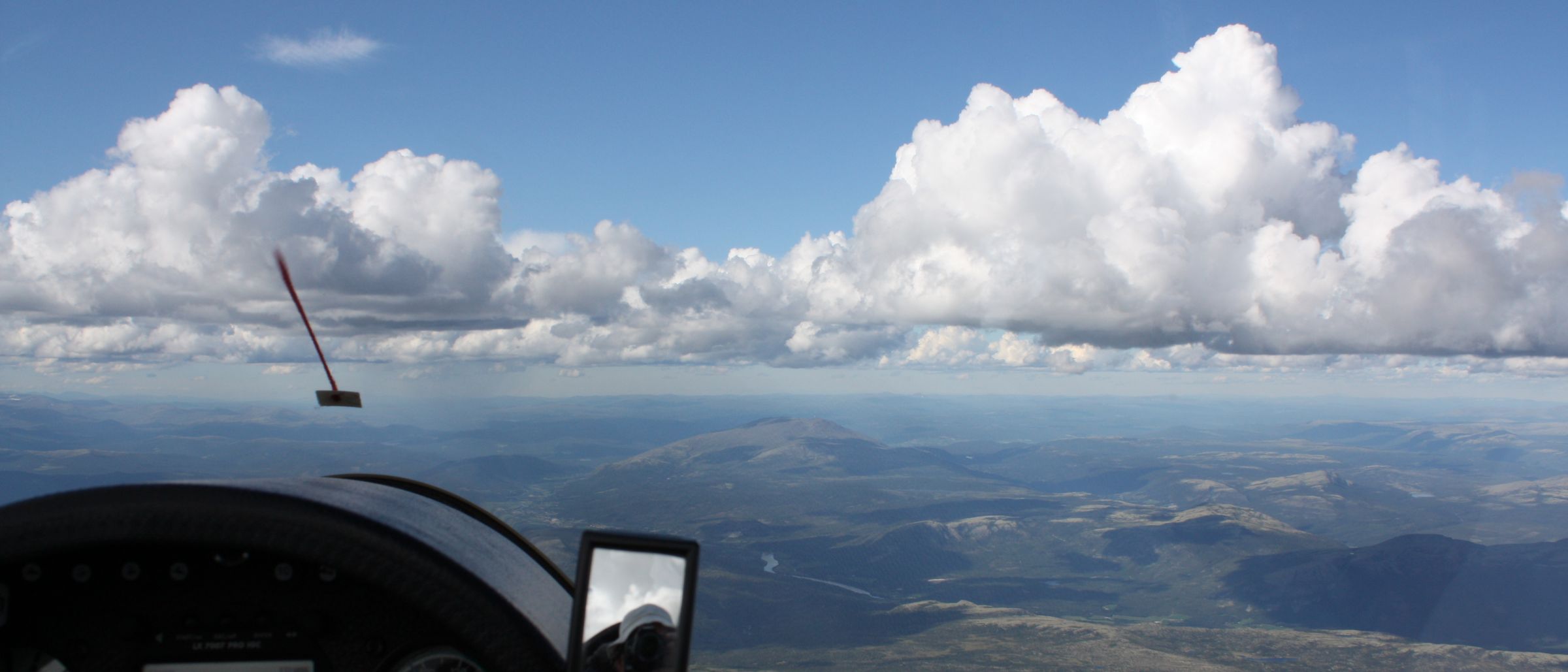 From a glider cockpit at height: the panel in shadow, cumulus along the horizon and Norwegian terrain below.